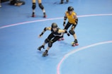 Two roller derby players, one in black and one in yellow, are skating on an indoor rink. Both are wearing protective gear including helmets and knee pads, and they appear to be in motion with a focus on speed. The blue floor has pink lines indicating the track.