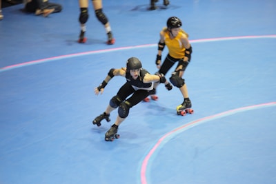 Two roller derby players, one in black and one in yellow, are skating on an indoor rink. Both are wearing protective gear including helmets and knee pads, and they appear to be in motion with a focus on speed. The blue floor has pink lines indicating the track.