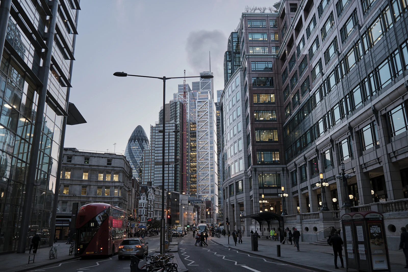 A dusk street view in the City of London, with the Gherkin skyscraper visible among office buildings and a red bus