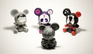 Four vinyl toy figures with mouse-like ears are arranged on a plain surface. The central figure is a gray toy with intricate white patterns, facing away. The others include a gray toy with a face mask design, a purple toy with a skull face, and a black toy with red accents and X marks on its eyes.