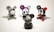 Four vinyl toy figures with mouse-like ears are arranged on a plain surface. The central figure is a gray toy with intricate white patterns, facing away. The others include a gray toy with a face mask design, a purple toy with a skull face, and a black toy with red accents and X marks on its eyes.