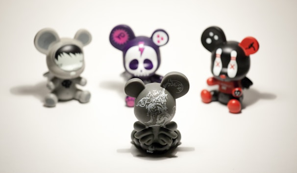 Four vinyl toy figures with mouse-like ears are arranged on a plain surface. The central figure is a gray toy with intricate white patterns, facing away. The others include a gray toy with a face mask design, a purple toy with a skull face, and a black toy with red accents and X marks on its eyes.