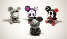 Four vinyl toy figures with mouse-like ears are arranged on a plain surface. The central figure is a gray toy with intricate white patterns, facing away. The others include a gray toy with a face mask design, a purple toy with a skull face, and a black toy with red accents and X marks on its eyes.