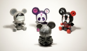 Four vinyl toy figures with mouse-like ears are arranged on a plain surface. The central figure is a gray toy with intricate white patterns, facing away. The others include a gray toy with a face mask design, a purple toy with a skull face, and a black toy with red accents and X marks on its eyes.