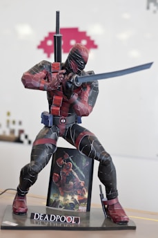 A detailed figure of a superhero in a crouched, dynamic pose holding a sword. The character wears a suit with a black and red color scheme and has accessories including a belt and holsters. The figure is mounted on a base labeled with a name. In the background, there is an additional image of the same character in an action-packed scene.