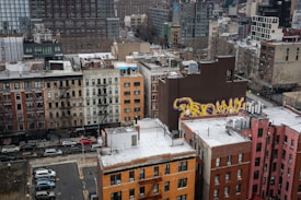 Aerial view of an urban landscape featuring a mix of residential and commercial buildings with varying architectural styles. A prominent brown building displays large graffiti art in yellow and purple. The scene is bustling with parked cars and streets lined with trees that are bare, indicating a colder season.