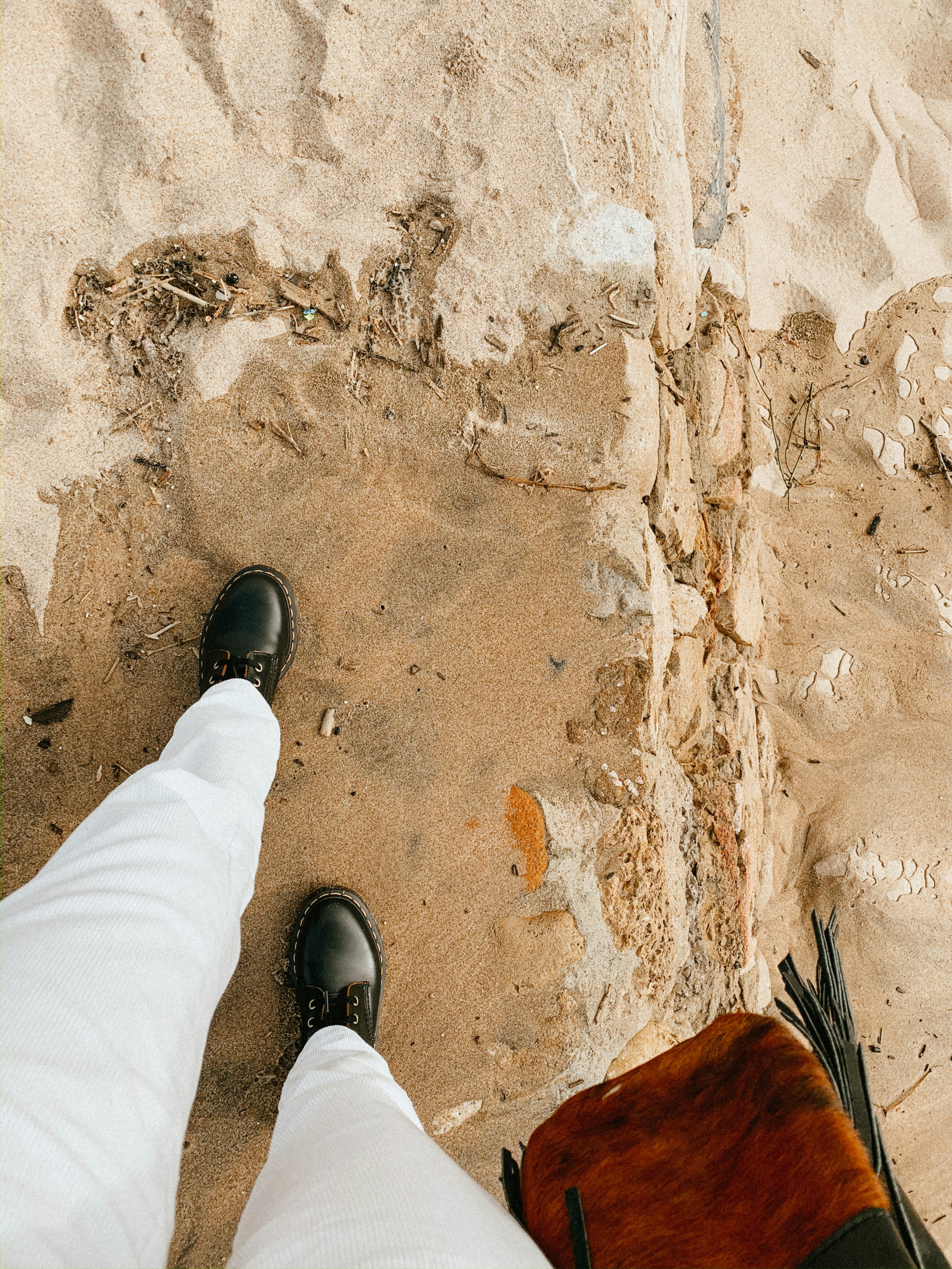 person in white pants and black leather shoes standing on brown sand
