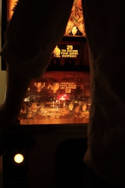 A dimly lit pinball machine with glowing orange and red lights, showing text and artwork. An individual is standing in front of the machine, with their arms partially visible, suggesting they are playing.