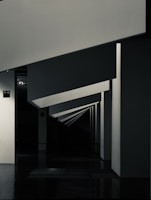 Geometric shapes and sharp angles create a modern architectural interior scene. The perspective of the image leads the eye through a series of receding plane segments, appearing as beams or dividers within the space. The lighting highlights the edges, adding depth to the darkened surroundings.