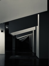 Geometric shapes and sharp angles create a modern architectural interior scene. The perspective of the image leads the eye through a series of receding plane segments, appearing as beams or dividers within the space. The lighting highlights the edges, adding depth to the darkened surroundings.