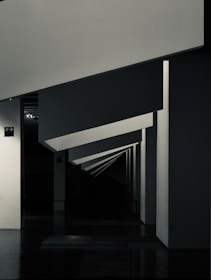 Geometric shapes and sharp angles create a modern architectural interior scene. The perspective of the image leads the eye through a series of receding plane segments, appearing as beams or dividers within the space. The lighting highlights the edges, adding depth to the darkened surroundings.