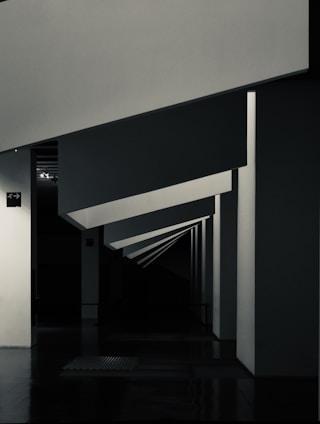 Geometric shapes and sharp angles create a modern architectural interior scene. The perspective of the image leads the eye through a series of receding plane segments, appearing as beams or dividers within the space. The lighting highlights the edges, adding depth to the darkened surroundings.