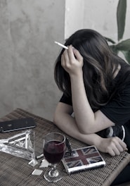 A person with long hair is sitting at a wicker table, holding a cigarette. On the table are a glass of red wine, a smartphone, a lighter, a book or notebook, and a few small reflective objects. The setting appears to be indoors with a neutral-toned wall and a plant in the background.