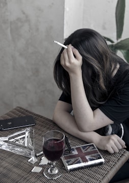 A person with long hair is sitting at a wicker table, holding a cigarette. On the table are a glass of red wine, a smartphone, a lighter, a book or notebook, and a few small reflective objects. The setting appears to be indoors with a neutral-toned wall and a plant in the background.