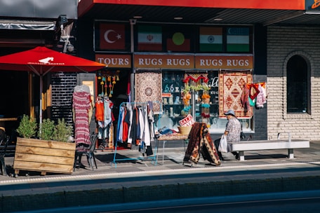 A street scene features a storefront with an array of colorful rugs, clothes, and decorative items displayed for sale. A person is sitting on a bench near the shop, and a bright red umbrella shades part of the outdoor area. Flags and a 'SALE' sign are also visible.