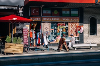 A street scene features a storefront with an array of colorful rugs, clothes, and decorative items displayed for sale. A person is sitting on a bench near the shop, and a bright red umbrella shades part of the outdoor area. Flags and a 'SALE' sign are also visible.