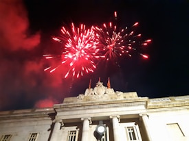Red fireworks burst in the dark night sky above a grand, ornately decorated building with visible flags. Smoke from the fireworks blends with the night sky, creating a dramatic effect.