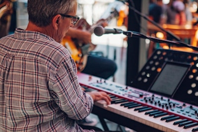 A musician wearing glasses and a plaid shirt plays a keyboard while sitting down. A microphone is set up in front while another musician can be partially seen playing a guitar in the background. The setting appears to be indoors with warm lighting, suggesting a live performance or band practice.