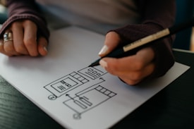 A person is drawing a flowchart on a piece of paper using a black pen. The flowchart includes various shapes and arrows indicating a process or sequence. The person is wearing a ring and long sleeve clothes with fingerless gloves.