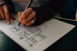 A person is sketching a wireframe or layout on a piece of paper with a black pen. The drawing includes several rectangles and lines that resemble website or app interface elements. The person's hand has a ring and their nails are painted white.