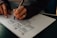 A person is sketching a wireframe or layout on a piece of paper with a black pen. The drawing includes several rectangles and lines that resemble website or app interface elements. The person's hand has a ring and their nails are painted white.