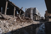 A street lined with partially destroyed buildings, debris, and rubble. One building on the left appears to be heavily damaged, with exposed rebar and a collapsed roof. The road is unpaved and features a large puddle reflecting the surroundings. The sky is clear and blue, casting shadows over the scene.