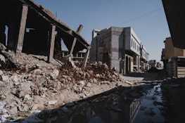 A street lined with partially destroyed buildings, debris, and rubble. One building on the left appears to be heavily damaged, with exposed rebar and a collapsed roof. The road is unpaved and features a large puddle reflecting the surroundings. The sky is clear and blue, casting shadows over the scene.