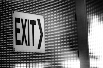 A black and white exit sign is affixed to a metal grid-like surface, featuring bold, uppercase letters directing towards the right.