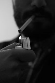 A person is holding a metal lighter with a small flame. The lighter is close to a cigarette in the person's mouth. The image is in black and white, creating a stark contrast between the lighter, the hand, and the partially visible face.