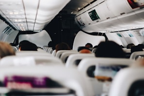 A view from inside a commercial airplane focusing on the cabin interior and passengers. Rows of seats are visible with overhead luggage compartments above. The lighting is artificial, creating a soft and comfortable atmosphere. Heads of passengers are visible, highlighting a sense of busyness and travel.