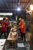 Several people are cooking at an outdoor food stall or small restaurant. They are using woks over open flames, with sparks flying in the air. The setting appears to be in an open-air structure with a tiled roof. A variety of kitchen utensils and cooking tools are visible, and the people are dressed casually in red shirts, indicating a possible uniform.