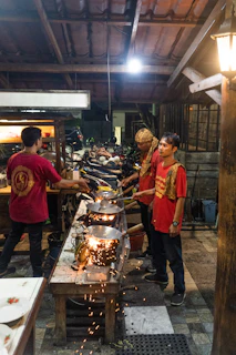 Students smiling as they stir woks filled with colorful ingredients over open flames.
