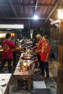 Several people are cooking at an outdoor food stall or small restaurant. They are using woks over open flames, with sparks flying in the air. The setting appears to be in an open-air structure with a tiled roof. A variety of kitchen utensils and cooking tools are visible, and the people are dressed casually in red shirts, indicating a possible uniform.