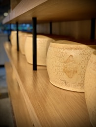 Several large wheels of cheese are lined up on wooden shelves. The cheese has a textured surface with engraved markings and text. The setting appears to be a storage or retail environment with natural wooden materials.