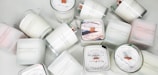 A collection of white candles, some with wooden wicks, arranged haphazardly on a flat surface. The candles are branded with 'French Candle Co' and have variations in size and shape. Some candles have lids with printed designs.