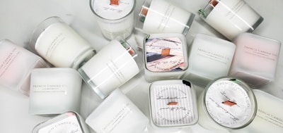A collection of white candles, some with wooden wicks, arranged haphazardly on a flat surface. The candles are branded with 'French Candle Co' and have variations in size and shape. Some candles have lids with printed designs.