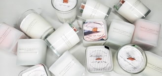 A collection of white candles, some with wooden wicks, arranged haphazardly on a flat surface. The candles are branded with 'French Candle Co' and have variations in size and shape. Some candles have lids with printed designs.