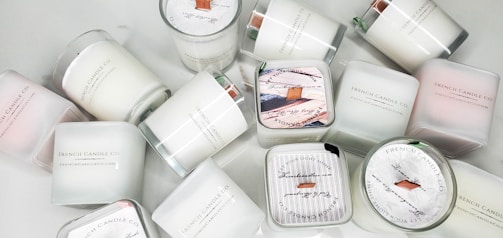 A collection of white candles, some with wooden wicks, arranged haphazardly on a flat surface. The candles are branded with 'French Candle Co' and have variations in size and shape. Some candles have lids with printed designs.