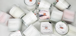 A collection of white candles, some with wooden wicks, arranged haphazardly on a flat surface. The candles are branded with 'French Candle Co' and have variations in size and shape. Some candles have lids with printed designs.