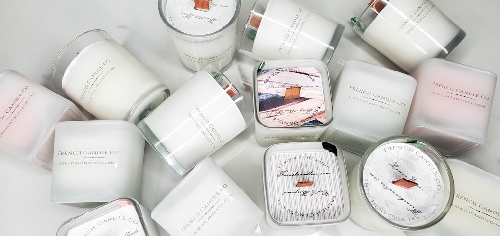 A collection of white candles, some with wooden wicks, arranged haphazardly on a flat surface. The candles are branded with 'French Candle Co' and have variations in size and shape. Some candles have lids with printed designs.