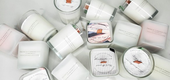 A collection of white candles, some with wooden wicks, arranged haphazardly on a flat surface. The candles are branded with 'French Candle Co' and have variations in size and shape. Some candles have lids with printed designs.