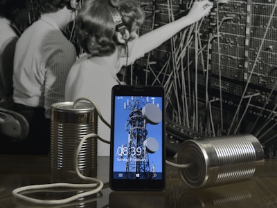 A black and white photograph of telephone operators working at a switchboard forms the background. In front, a modern smartphone displays an image of a communication tower. Connected to the phone are two tin cans with a string, reminiscent of a makeshift phone.