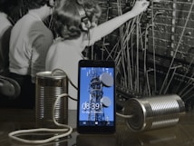 A black and white photograph of telephone operators working at a switchboard forms the background. In front, a modern smartphone displays an image of a communication tower. Connected to the phone are two tin cans with a string, reminiscent of a makeshift phone.
