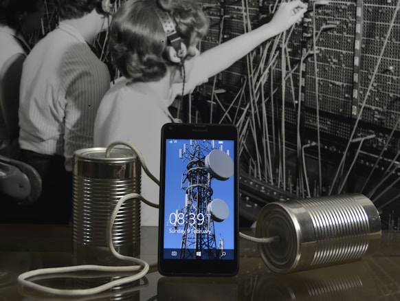 A black and white photograph of telephone operators working at a switchboard forms the background. In front, a modern smartphone displays an image of a communication tower. Connected to the phone are two tin cans with a string, reminiscent of a makeshift phone.