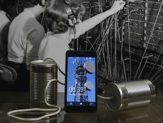 A black and white photograph of telephone operators working at a switchboard forms the background. In front, a modern smartphone displays an image of a communication tower. Connected to the phone are two tin cans with a string, reminiscent of a makeshift phone.