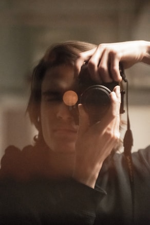 A person holding a camera up to their face, capturing a close-up reflection or self-portrait in a mirror. The image has a soft focus with warm lighting, emphasizing the intimate and personal nature of the scene.