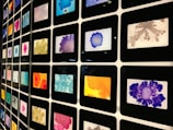 A grid of framed colorful abstract art pieces arranged in a matrix formation. Each frame contains unique patterns with various vibrant colors including blue, orange, purple, red, pink, and gray. The art pieces appear to be inspired by ink or watercolor splatters and blobs.