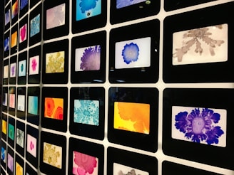 A grid of framed colorful abstract art pieces arranged in a matrix formation. Each frame contains unique patterns with various vibrant colors including blue, orange, purple, red, pink, and gray. The art pieces appear to be inspired by ink or watercolor splatters and blobs.