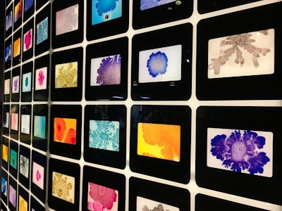 A grid of framed colorful abstract art pieces arranged in a matrix formation. Each frame contains unique patterns with various vibrant colors including blue, orange, purple, red, pink, and gray. The art pieces appear to be inspired by ink or watercolor splatters and blobs.