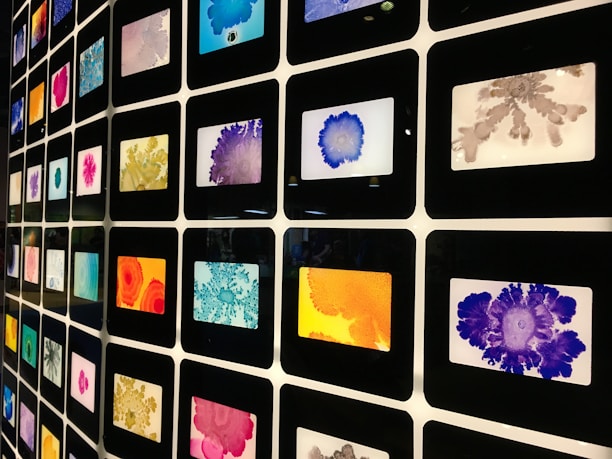 A grid of framed colorful abstract art pieces arranged in a matrix formation. Each frame contains unique patterns with various vibrant colors including blue, orange, purple, red, pink, and gray. The art pieces appear to be inspired by ink or watercolor splatters and blobs.